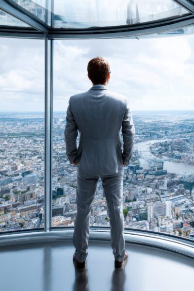 Man looking out window A man in a suit admires a sprawling urban landscape from a glass-walled observation deck.